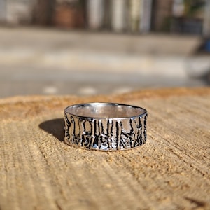 May include: A silver-colored ring with a textured, bark-like design. The ring is circular and sits on a wooden surface, with the sunlight casting a shadow. The ring's surface has a detailed, irregular pattern.
