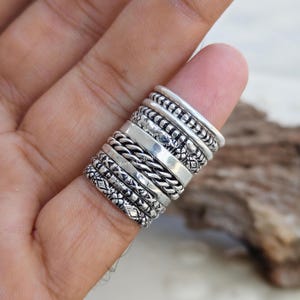 Puede incluir: Una pila de diez anillos de plata en un dedo. Los anillos presentan varios diseños, incluyendo bandas lisas, patrones de cuerda trenzada y bandas con motivos geométricos y florales grabados. El metal tiene un acabado pulido con áreas oscurecidas.