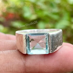 May include: A silver ring featuring a square-cut, light blue gemstone set in the center. The band has a smooth, polished finish on the sides. The text SilverStudio26 is visible above the ring. The design is simple and focuses on the stone.