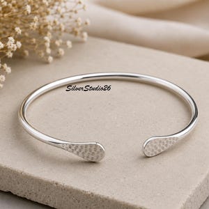 Peut inclure: Un bracelet manchette en argent avec un anneau lisse et poli et deux extrémités aplaties à la texture martelée. La surface métallique est réfléchissante et brillante. Le texte SilverStudio26 est imprimé en noir au centre du bracelet ouvert.