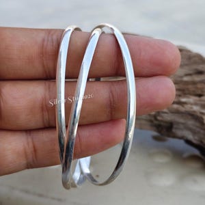 Könnte beinhalten: Ein Set aus zwei ineinandergreifenden Silberarmreifen mit einer glatten, polierten und reflektierenden metallischen Oberfläche. Die kreisförmigen Armreifen haben ein schlichtes, abgerundetes Profil und zeigen eine helle, saubere und gleichmäßige Silberoptik.