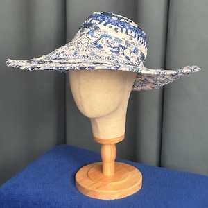 May include: A wide-brimmed sun hat featuring a blue and white patterned fabric with floral and geometric motifs. The hat has a structured crown and a broad, slightly wavy brim, displaying a quilted texture throughout the material.