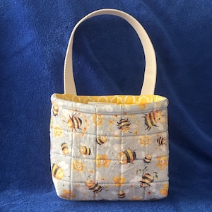 May include: A quilted fabric basket featuring a light gray background with a repeating pattern of yellow and black bees and smiling yellow flowers. The basket has a single cream-colored handle and a bright yellow interior lining with a white pattern.