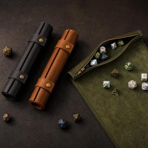 Puede incluir: Dos estuches cilíndricos de cuero en negro y marrón, cada uno asegurado con un botón de presión de latón. Junto a ellos hay una bandeja para dados de ante verde oliva abierta con un compartimento con cremallera y varios dados poliédricos.
