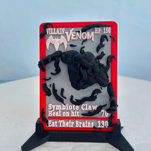 Puede incluir: Una tarjeta rectangular impresa en 3D con una figura negra de Venom sobre un fondo gris con borde rojo. El texto dice Villain, Venom, HP 150, Symbiote Claw, Heal on hit 70 y Eat Their Brains 130. La tarjeta se sostiene sobre una base negra.