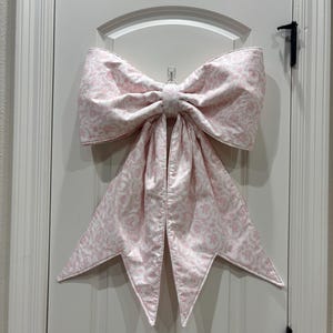 May include: A large decorative bow made of fabric with a light pink and white floral toile pattern. The bow features two wide, structured loops at the top and two long, pointed tails hanging downward, suspended by a clear plastic hook.