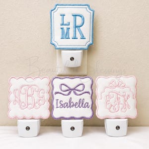 May include: Four square night lights with white fabric covers featuring scalloped edges and embroidered designs. One has blue monogram letters LMR, one has pink monogram letters ABG, one has a purple bow and the name Isabella, and one has pink monogram letters FK.