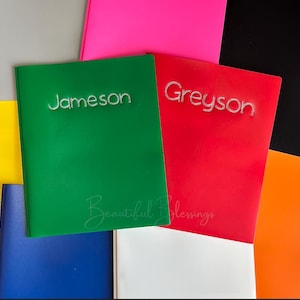 May include: A green notebook with the name Jameson in silver rhinestones and a red notebook with the name Greyson in silver rhinestones, surrounded by various colorful notebooks in blue, white, orange, pink, black, and yellow.