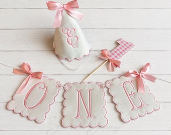 May include: A white party hat with a pink satin bow and embroidered monogram, a pink and white gingham number one cake topper on a wooden stick, and a white scalloped banner with pink embroidered letters spelling out the word ONE.