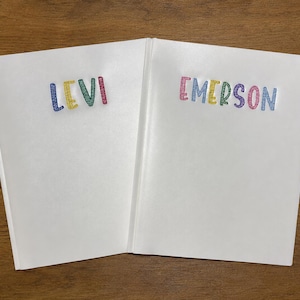 May include: Two white hardcover books with personalized names embroidered on the front covers. The left book features the name LEVI in multicolored stitched letters, and the right book features the name EMERSON in multicolored stitched letters.