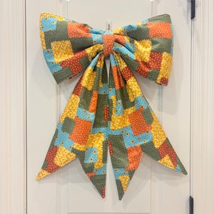 May include: A large fabric bow featuring a patchwork design with various patterns in shades of orange, yellow, light blue, and olive green. The bow has two wide loops and two long, pointed tails hanging down, all constructed from quilted fabric.
