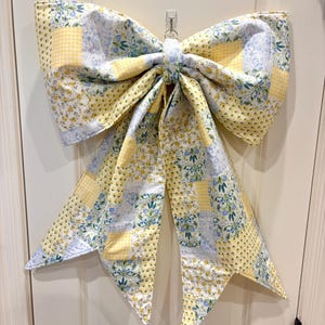 May include: A large decorative bow made of quilted fabric featuring a patchwork pattern of yellow gingham, light blue floral prints, and white sections with small yellow and blue flowers. The bow has two wide loops and two long, pointed tails.