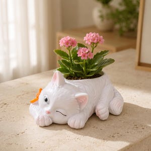 May include: A white cat-shaped planter featuring a textured fur design, a small orange bow on the side, and a winking expression. The planter holds a small green plant with clusters of vibrant pink flowers blooming from the top of the cat's back.