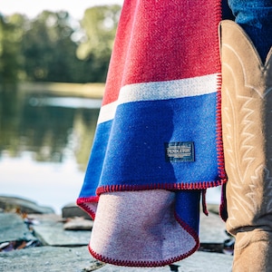 May include: A red, white, and blue Pendleton blanket with a red stitched edge. The blanket is draped over a person's leg, with a cowboy boot visible. The blanket has a label that reads "Pendleton".