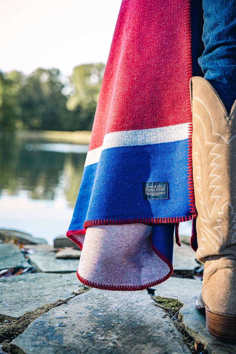 May include: A red, white, and blue Pendleton blanket with a red stitched edge. The blanket is draped over a person's leg, with a cowboy boot visible. The blanket has a label that reads "Pendleton".