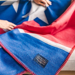 May include: A blue, red, and white blanket with a star design. The blanket has a red stitched edge and a rectangular label that reads "Pendleton". The blanket is folded over a gray couch.
