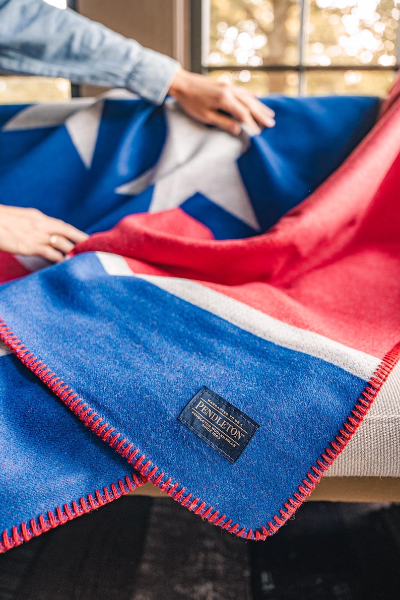 May include: A blue, red, and white blanket with a star design. The blanket has a red stitched edge and a rectangular label that reads "Pendleton". The blanket is folded over a gray couch.