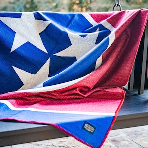 May include: A blue, red, and white blanket with a star pattern. The blanket is draped over a dark surface, with the stars in white against a blue background. The blanket has a red border and a tag.