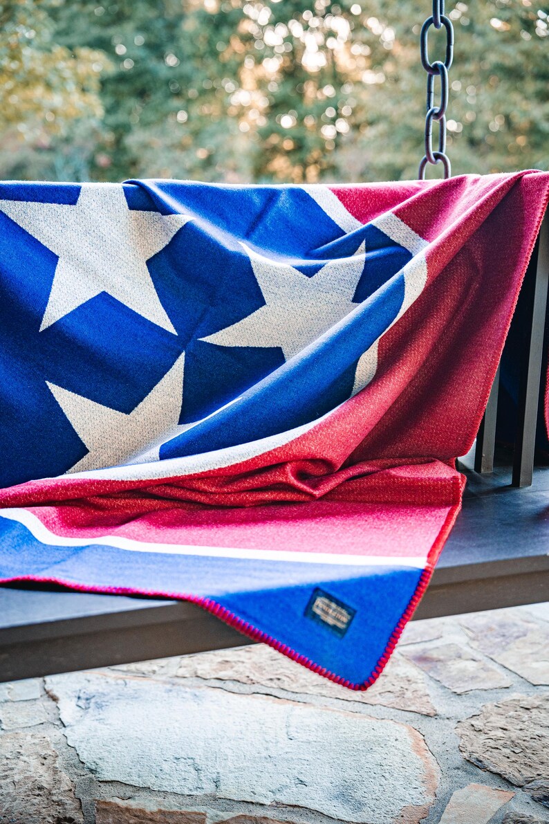 May include: A blue, red, and white blanket with a star pattern. The blanket is draped over a dark surface, with the stars in white against a blue background. The blanket has a red border and a tag.