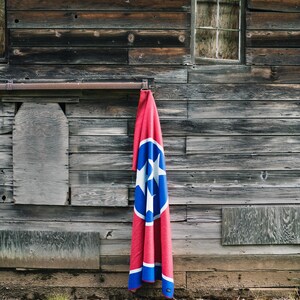 May include: A red and blue blanket with the Tennessee state flag design hanging on a weathered wooden wall. The blanket features a large blue circle with three white stars and a white border.