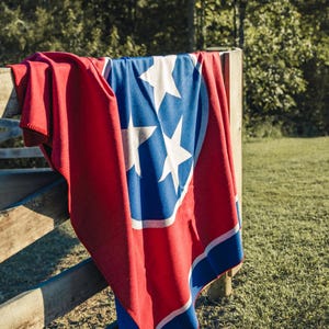 May include: A red, white, and blue blanket draped over a wooden fence. The blanket features the Tennessee state flag design, with three white stars inside a blue circle, set against a red background.