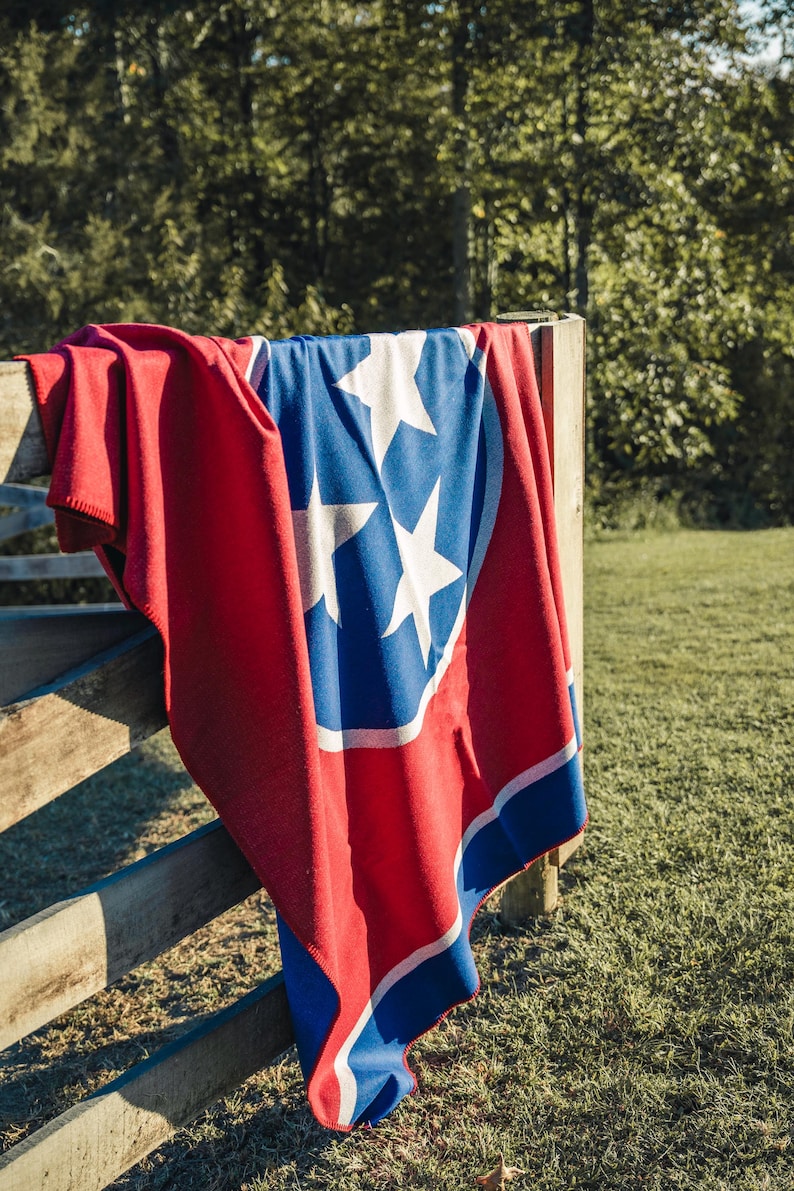 May include: A red, white, and blue blanket draped over a wooden fence. The blanket features the Tennessee state flag design, with three white stars inside a blue circle, set against a red background.