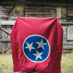 May include: A red blanket with the Tennessee state flag design. The flag features a blue circle with three white stars in the center, and a blue stripe at the bottom. The blanket is displayed outdoors.