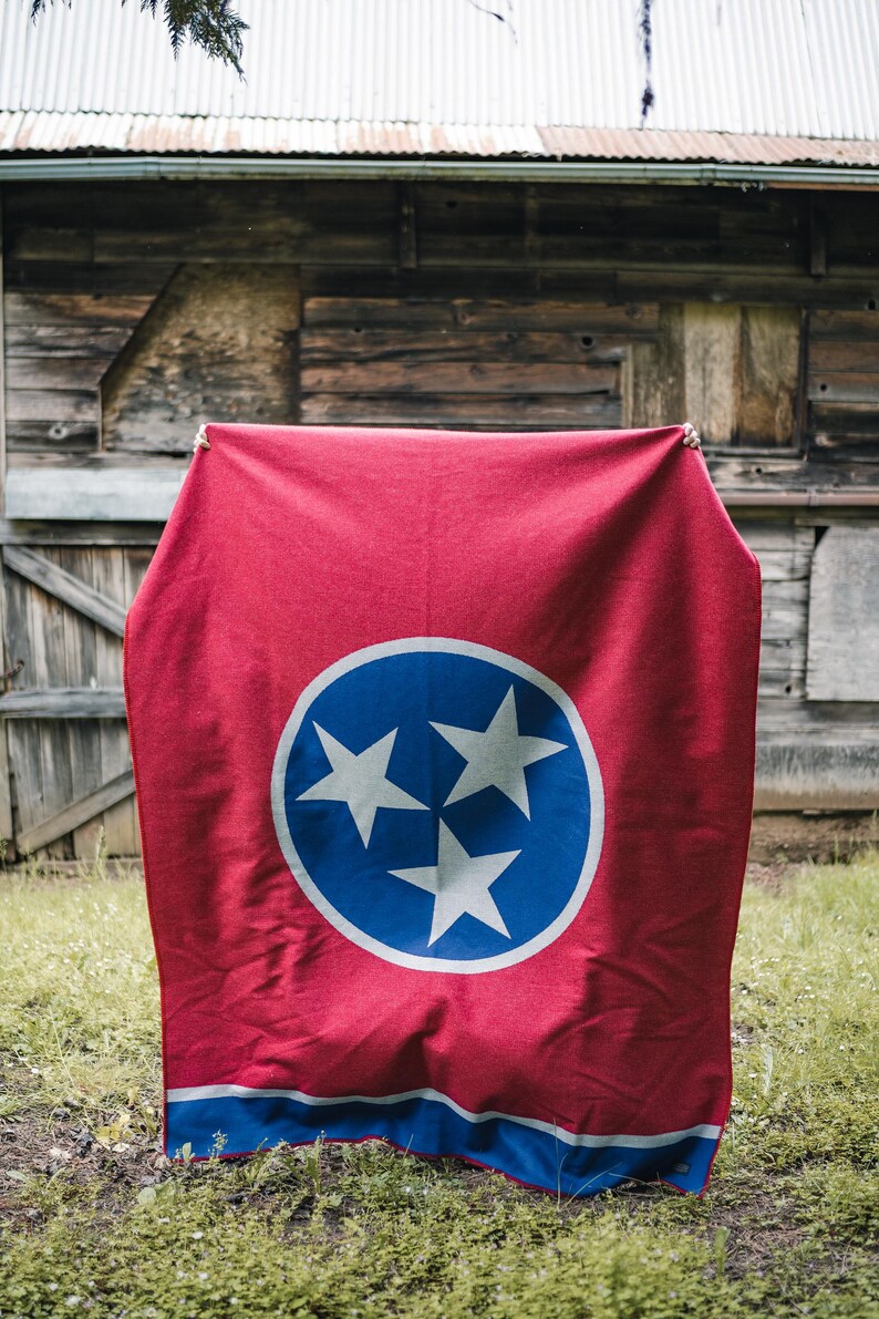 May include: A red blanket with the Tennessee state flag design. The flag features a blue circle with three white stars in the center, and a blue stripe at the bottom. The blanket is displayed outdoors.