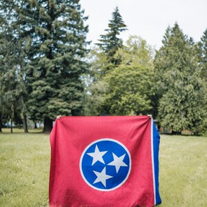 May include: A red and blue flag featuring the Tennessee state emblem, a white circle with three white stars inside a blue circle. The flag is displayed outdoors on a grassy field with trees in the background.