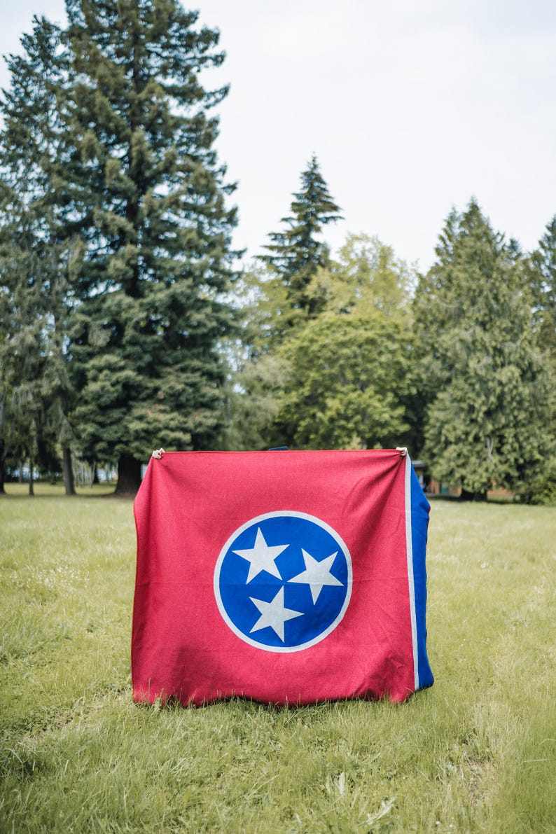 May include: A red and blue flag featuring the Tennessee state emblem, a white circle with three white stars inside a blue circle. The flag is displayed outdoors on a grassy field with trees in the background.