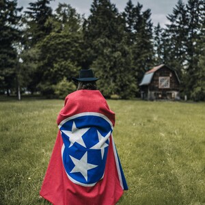 May include: A red and blue cape featuring the Tennessee state flag design, worn by a person in a black hat, stands out against a green field and forest backdrop. The cape's design includes a blue circle with three white stars and a white border.