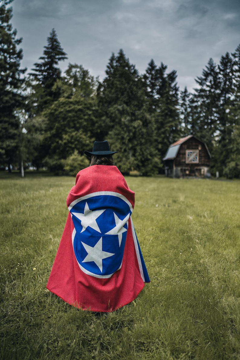 May include: A red and blue cape featuring the Tennessee state flag design, worn by a person in a black hat, stands out against a green field and forest backdrop. The cape's design includes a blue circle with three white stars and a white border.