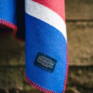 May include: A folded blanket with a blue, red, and white design. The blanket has a blue field with a red stitched edge. A label with the brand name "Pendleton" is visible.