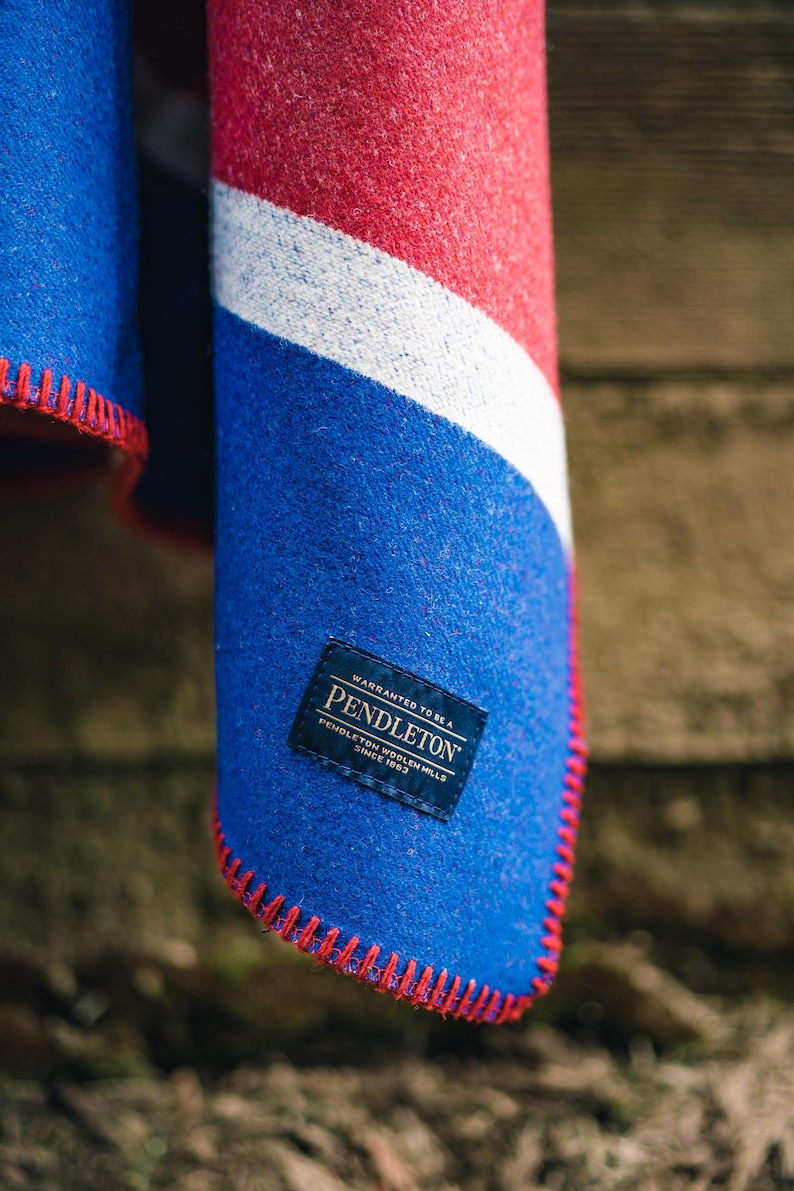 May include: A folded blanket with a blue, red, and white design. The blanket has a blue field with a red stitched edge. A label with the brand name "Pendleton" is visible.