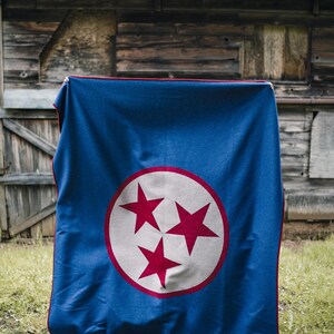 May include: A blue blanket with a red and white design. The blanket features a large white circle with three red stars inside. The border of the blanket is red. The blanket is hanging outside on a metal rod.