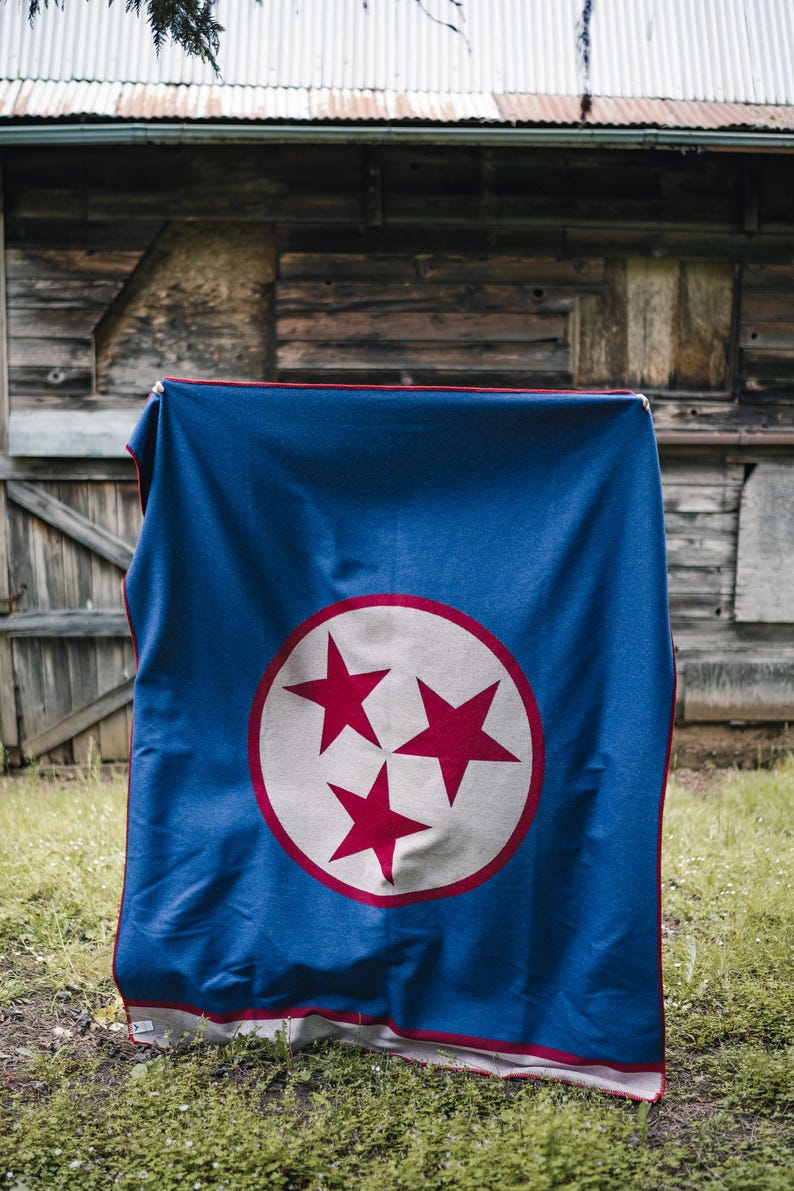 May include: A blue blanket with a red and white design. The blanket features a large white circle with three red stars inside. The border of the blanket is red. The blanket is hanging outside on a metal rod.
