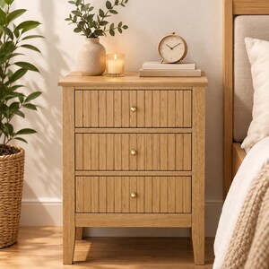 May include: A light oak wood nightstand featuring three drawers with vertical fluted detailing and small round brass knobs. The nightstand has a rectangular top surface and four straight legs, providing a stable base for bedroom storage.