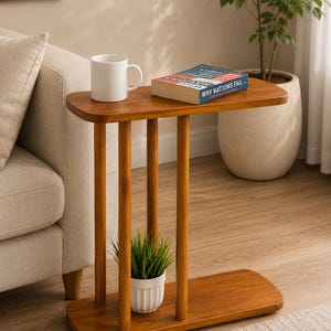 May include: A C-shaped side table crafted from warm-toned wood, featuring a smooth rectangular top and a matching base connected by three vertical cylindrical supports. A white mug and a book titled Why Nations Fail sit on the table surface.