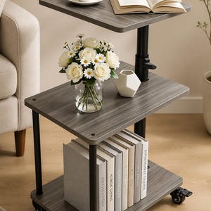 May include: A three-tiered rolling side table with a dark gray wood grain finish and black metal supports. The table features a height-adjustable top shelf, a middle shelf holding a glass vase with white roses and daisies, and a bottom shelf with books.
