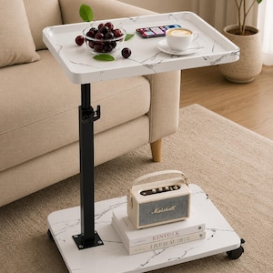May include: A white marble-patterned rolling side table with a black adjustable metal post. The top surface holds a glass bowl of cherries, a smartphone, and a coffee cup. The base shelf holds a beige Marshall speaker and two stacked books.