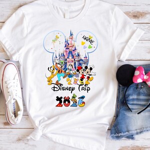 May include: A white t-shirt featuring a colorful graphic of a castle and various cartoon characters, with the text "NAME", "Disney Trip", and "2026" printed on the front. The shirt is displayed alongside white sneakers and denim shorts.