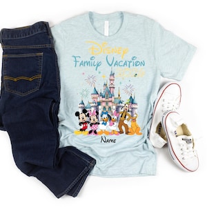 May include: A light blue heathered t-shirt featuring a graphic of a castle, fireworks, and Disney characters including Mickey, Minnie, Donald, Daisy, Goofy, and Pluto. The text reads Disney Family Vacation 2026 and Name. Dark blue jeans and white sneakers.