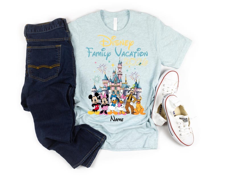 May include: A light blue heathered t-shirt featuring a graphic of a castle, fireworks, and Disney characters including Mickey, Minnie, Donald, Daisy, Goofy, and Pluto. The text reads Disney Family Vacation 2026 and Name. Dark blue jeans and white sneakers.