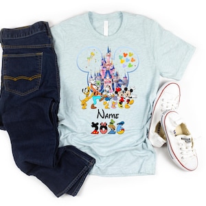 K&ouml;nnte beinhalten: Ein hellblaues, meliertes T-Shirt mit einer Grafik eines Schlosses in einer Mickey-Mouse-Silhouette, umgeben von Zeichentrickfiguren, dem Text Name und der Jahreszahl 2026. Dunkelblaue Jeans und wei&szlig;e Stoffschuhe liegen daneben.