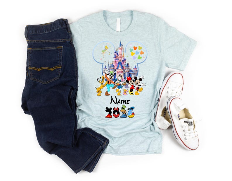 K&ouml;nnte beinhalten: Ein hellblaues, meliertes T-Shirt mit einer Grafik eines Schlosses in einer Mickey-Mouse-Silhouette, umgeben von Zeichentrickfiguren, dem Text Name und der Jahreszahl 2026. Dunkelblaue Jeans und wei&szlig;e Stoffschuhe liegen daneben.