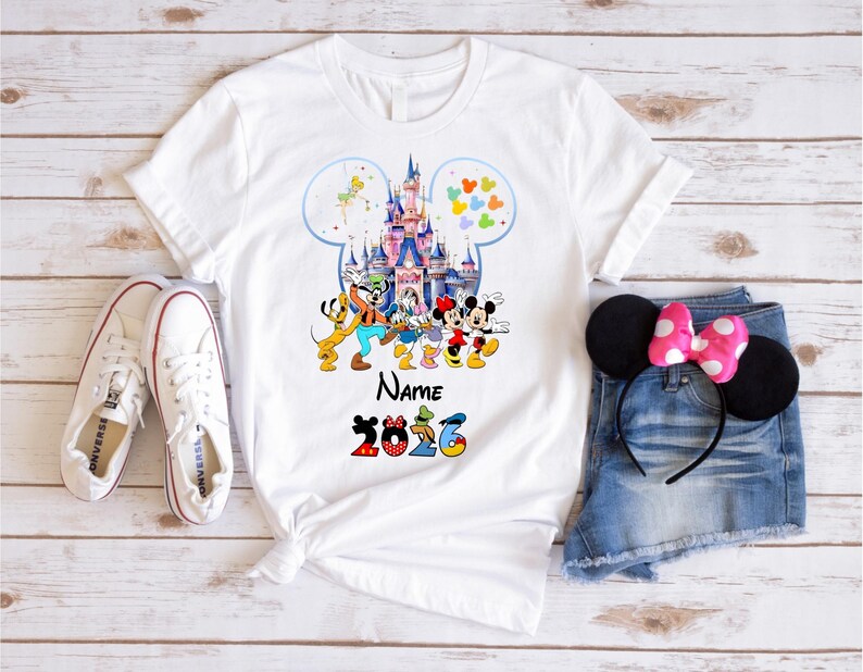May include: A white short-sleeved t-shirt featuring a graphic of a castle inside a Mickey Mouse silhouette, surrounded by cartoon characters, the word Name, and the year 2026. The shirt is displayed with white sneakers and denim shorts.