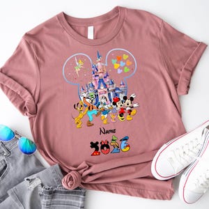 K&ouml;nnte beinhalten: Ein altrosa T-Shirt mit einer Grafik eines Schlosses in einer Micky-Maus-Ohren-Silhouette, umgeben von Zeichentrickfiguren wie Micky, Minnie, Donald, Daisy, Goofy, Pluto und Tinkerbell, mit dem Text Name und 2026 darunter.