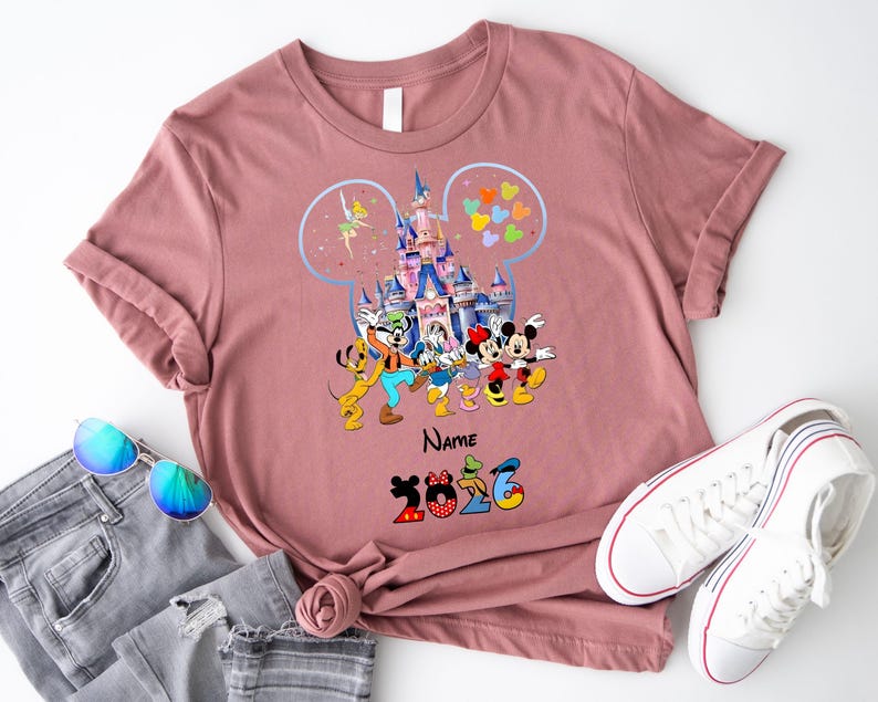 K&ouml;nnte beinhalten: Ein altrosa T-Shirt mit einer Grafik eines Schlosses in einer Micky-Maus-Ohren-Silhouette, umgeben von Zeichentrickfiguren wie Micky, Minnie, Donald, Daisy, Goofy, Pluto und Tinkerbell, mit dem Text Name und 2026 darunter.