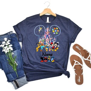 K&ouml;nnte beinhalten: Ein marineblaues Kurzarm-T-Shirt mit einer Grafik eines Schlosses in einer Micky-Maus-Ohr-Silhouette, umgeben von Zeichentrickfiguren, dem Wort Name und der Zahl 2026. Das T-Shirt wird zusammen mit einer blauen Jeans und Sandalen gezeigt.