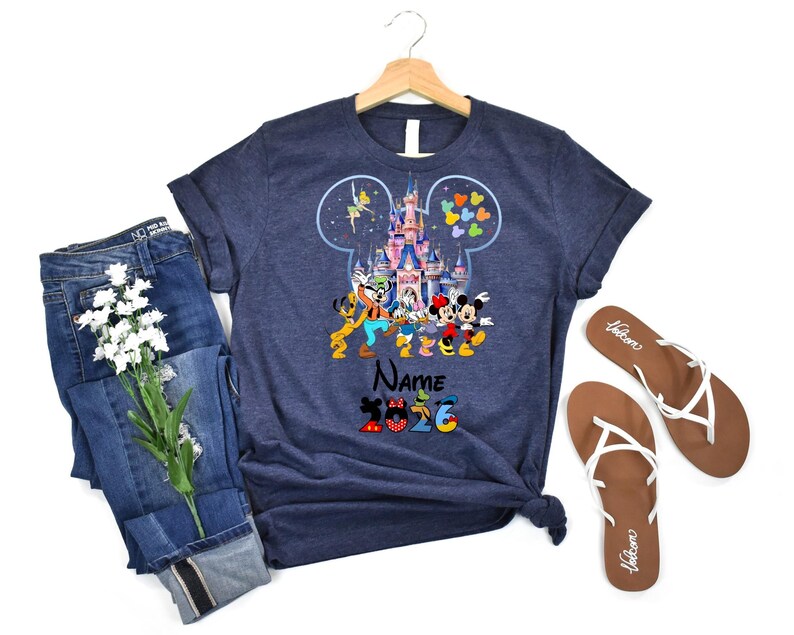 K&ouml;nnte beinhalten: Ein marineblaues Kurzarm-T-Shirt mit einer Grafik eines Schlosses in einer Micky-Maus-Ohr-Silhouette, umgeben von Zeichentrickfiguren, dem Wort Name und der Zahl 2026. Das T-Shirt wird zusammen mit einer blauen Jeans und Sandalen gezeigt.